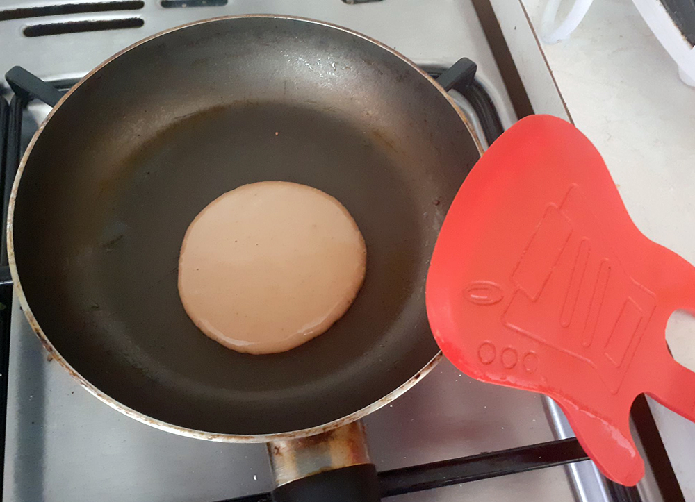 A very tiny pancake in a small pan next to a massive red spatula